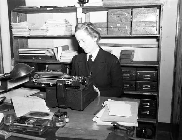 Black and white photograph. A woman with styled hair sits at a desk, typing on a typewriter. Behind her are shelves filled with paperwork and boxes. There are additional papers on the desk, as well as a small lamp.